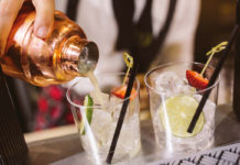 a cocktail is poured from a copper shaker into two gin glasses