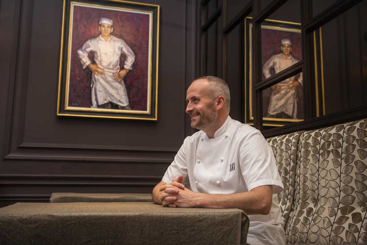 Head-Chef-Stevie-McLaughlin sits under a painting in chef whites