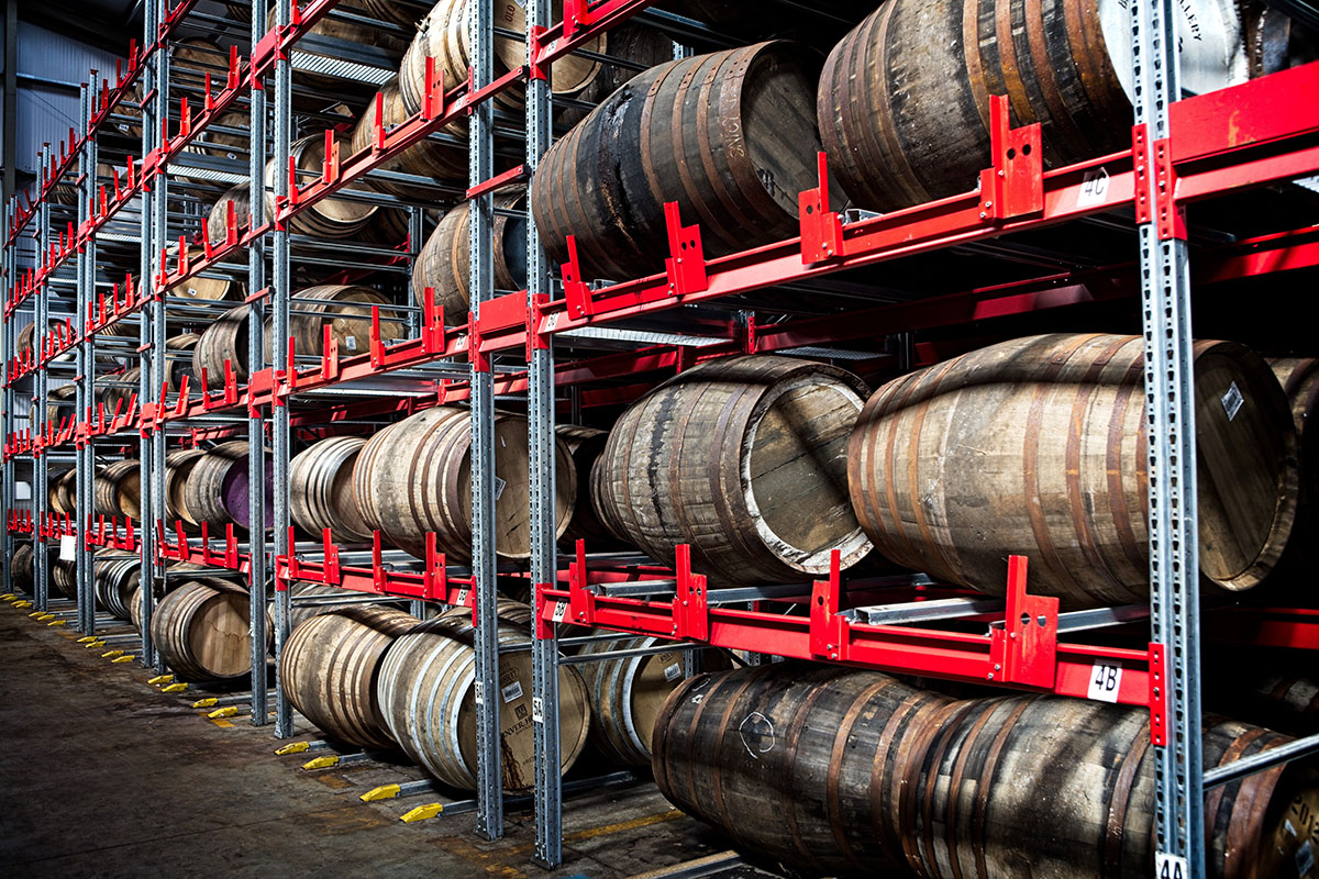 Photograph of multiple barrels stored on shelves in a distillery. 