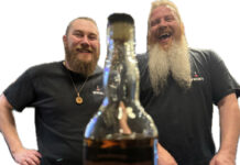 two bearded men wearing black t-shirts smile behind a bottle of whisky