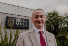 a man wearing a grey blazer, white shirt and red tie stands in front of the isle of mull hotel