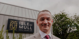 a man wearing a grey blazer, white shirt and red tie stands in front of the isle of mull hotel