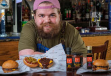 a chef wearing a pink cap and tartan apron stands with burgers and sauces