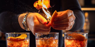 a bartended lights an orange peel garnish while making three old fashioned cocktails