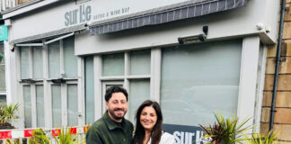 a man wearing a green coat and a woman in a white coat stand in front of a wine bar