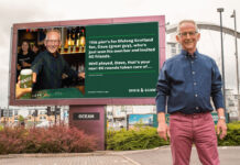 A middle-aged man stands in front of a billboard with his own face on it