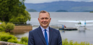 a man in a suit stands in front of a loch where a sea-plane is moored