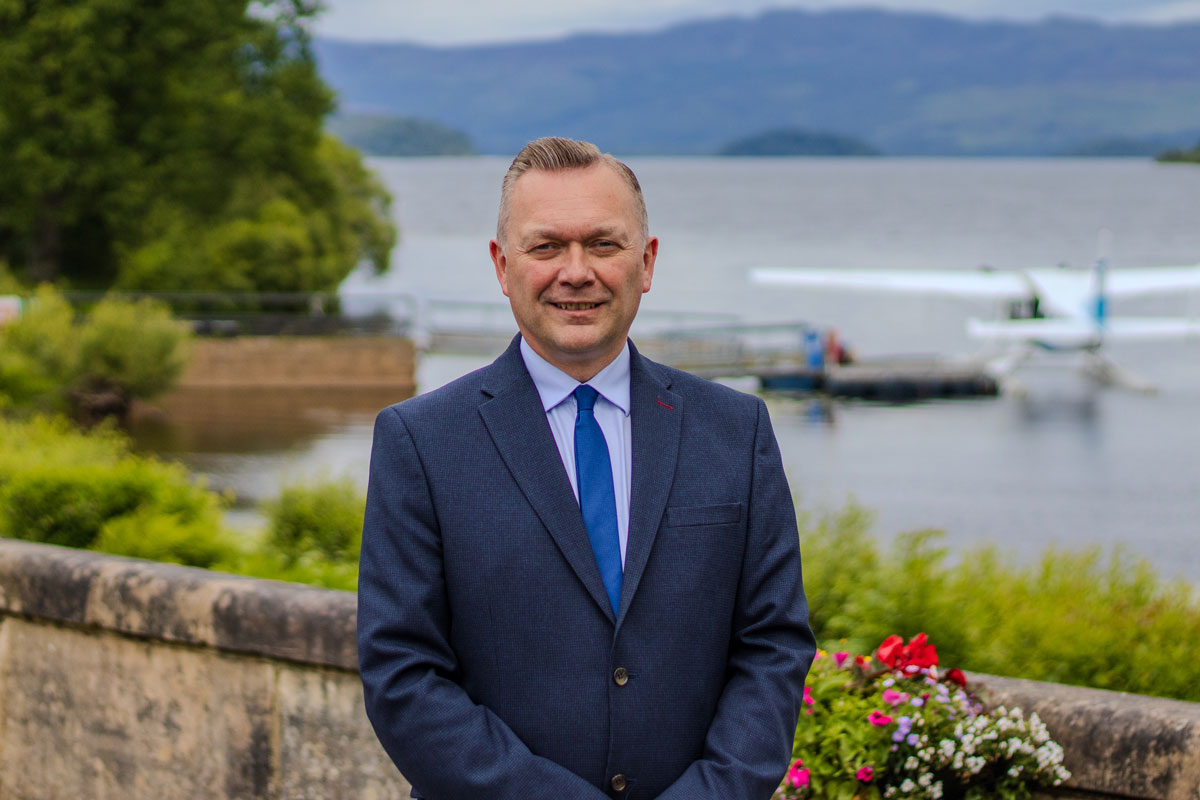 a man in a suit stands in front of a loch where a sea-plane is moored