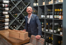 A smartly dressed man smiles in front of a large wine rack