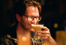 A smiling bespectacled young man drinks a cocktail