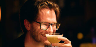 A smiling bespectacled young man drinks a cocktail