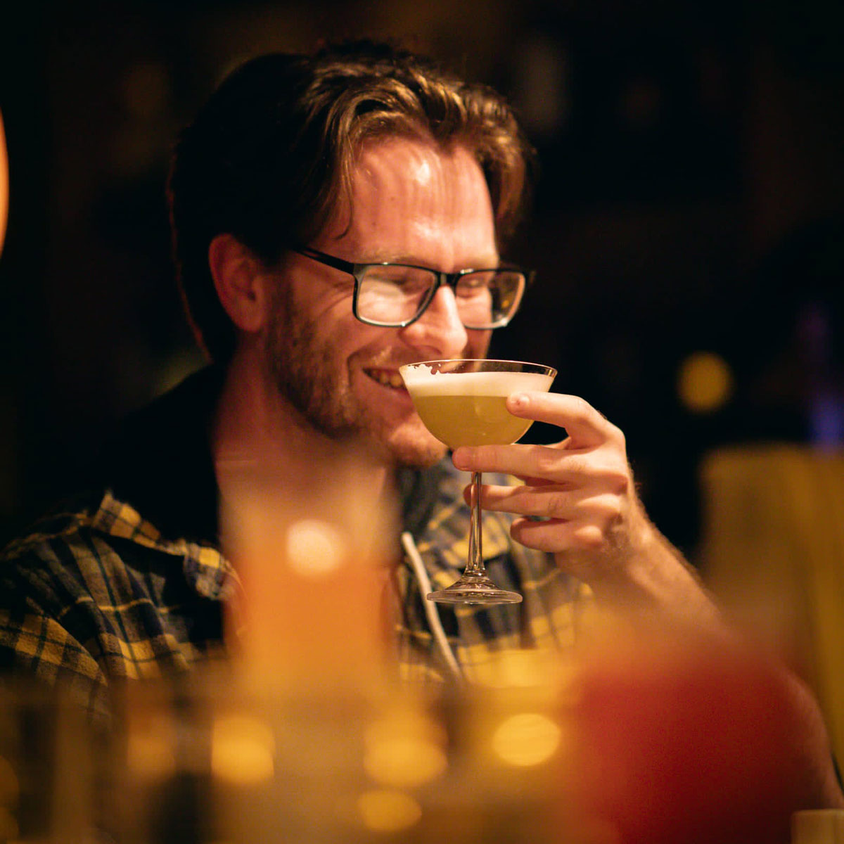 A smiling bespectacled young man drinks a cocktail