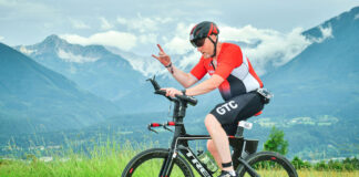 A man in red lycra cycles along an Alpine road, giving the hand signal for 'heavy metal'