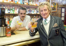 rod stewart holds a whisky cocktail at a bar next to a bottle of his wolfies whisky