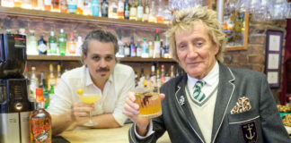 rod stewart holds a whisky cocktail at a bar next to a bottle of his wolfies whisky