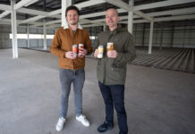 Two men stand in an industrial space holding cans of beer