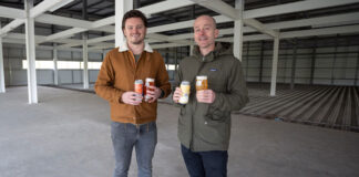 Two men stand in an industrial space holding cans of beer