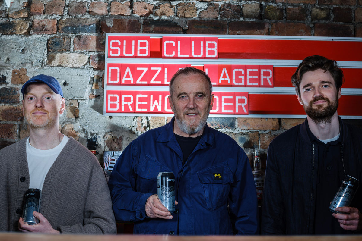 Three men stand indoors with cans of lager