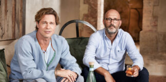 Two casually dressed men sit at a low table with bottle on it