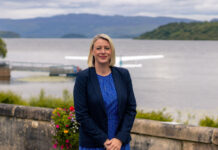 A well-dressed woman stands beside a loch, with a seaplane behind her