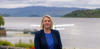 A well-dressed woman stands beside a loch, with a seaplane behind her
