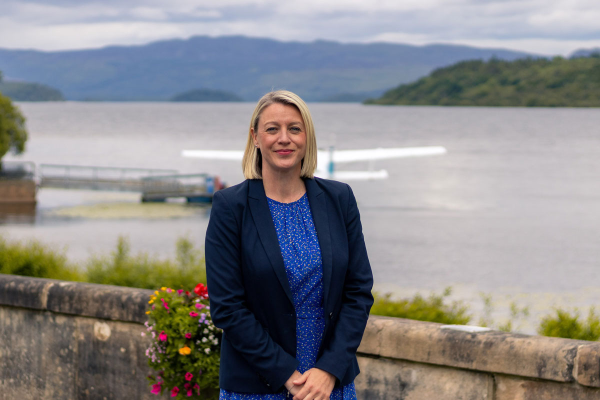 A well-dressed woman stands beside a loch, with a seaplane behind her