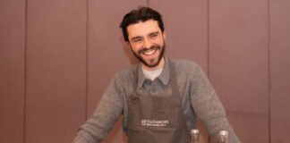 A young bearded man grins behind a bar