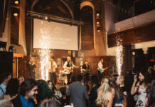 a room full of young people dance to a band onstage as fireworks go off