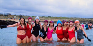 A group of women in bright bathing costumes standing in the sea with the beach behind them