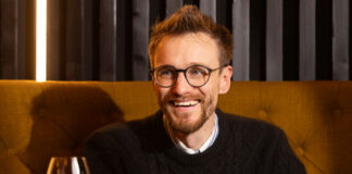 a young bearded and bespectacled man laughs over a drink