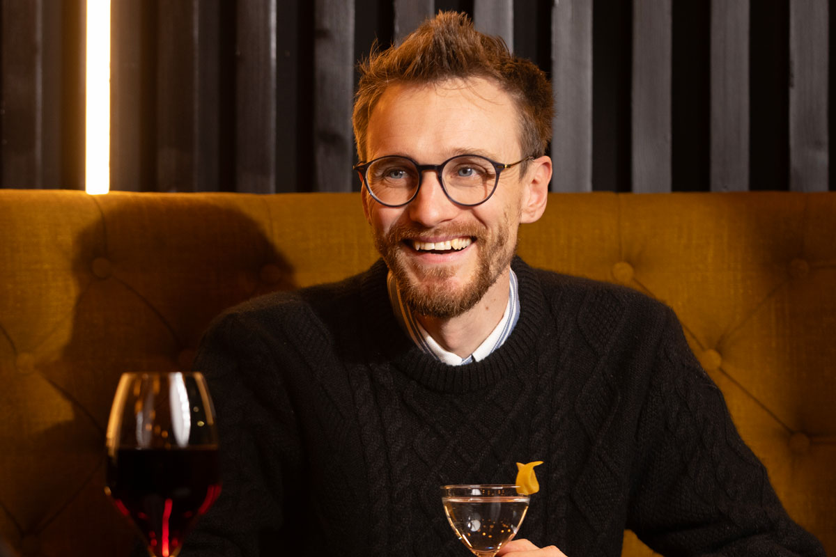 a young bearded and bespectacled man laughs over a drink