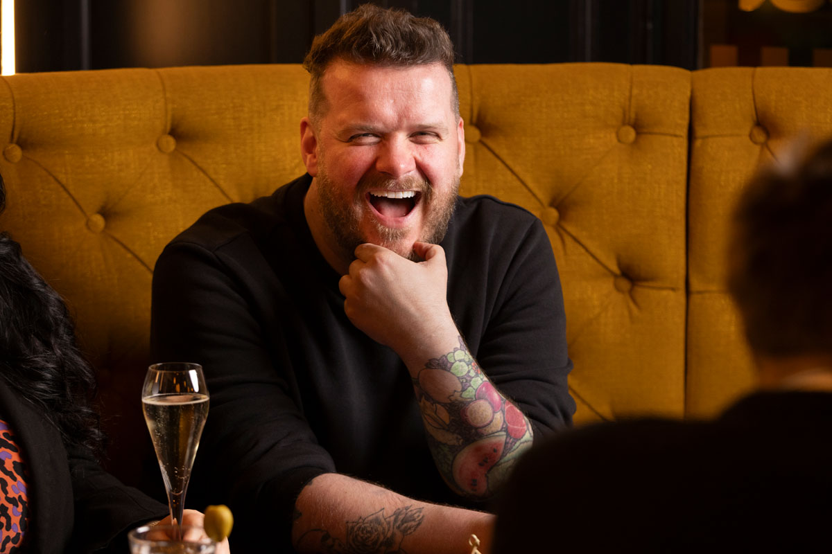 A bearded man laughs whilst sitting in a bar environment
