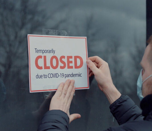 Man wearing medical mask puts a Temporary closed due COVID-19 pandemic sign on a window