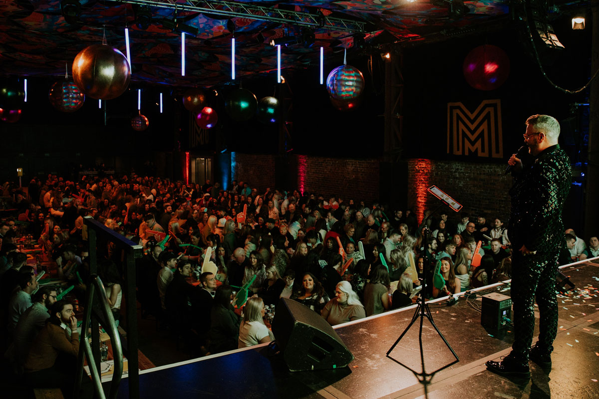 A man stands on stage in front of a busy room of revellers
