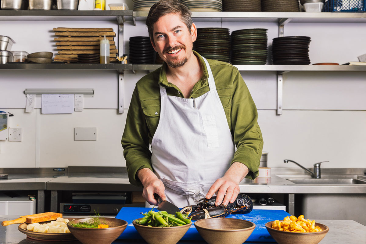 A smiling and relaxed bearded man, wearing an apron, prepares food in a kitchen.