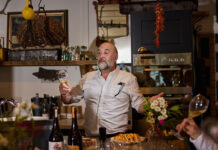 a jolly-looking bearded man holds forth over a table of food and wine