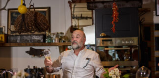 a jolly-looking bearded man holds forth over a table of food and wine