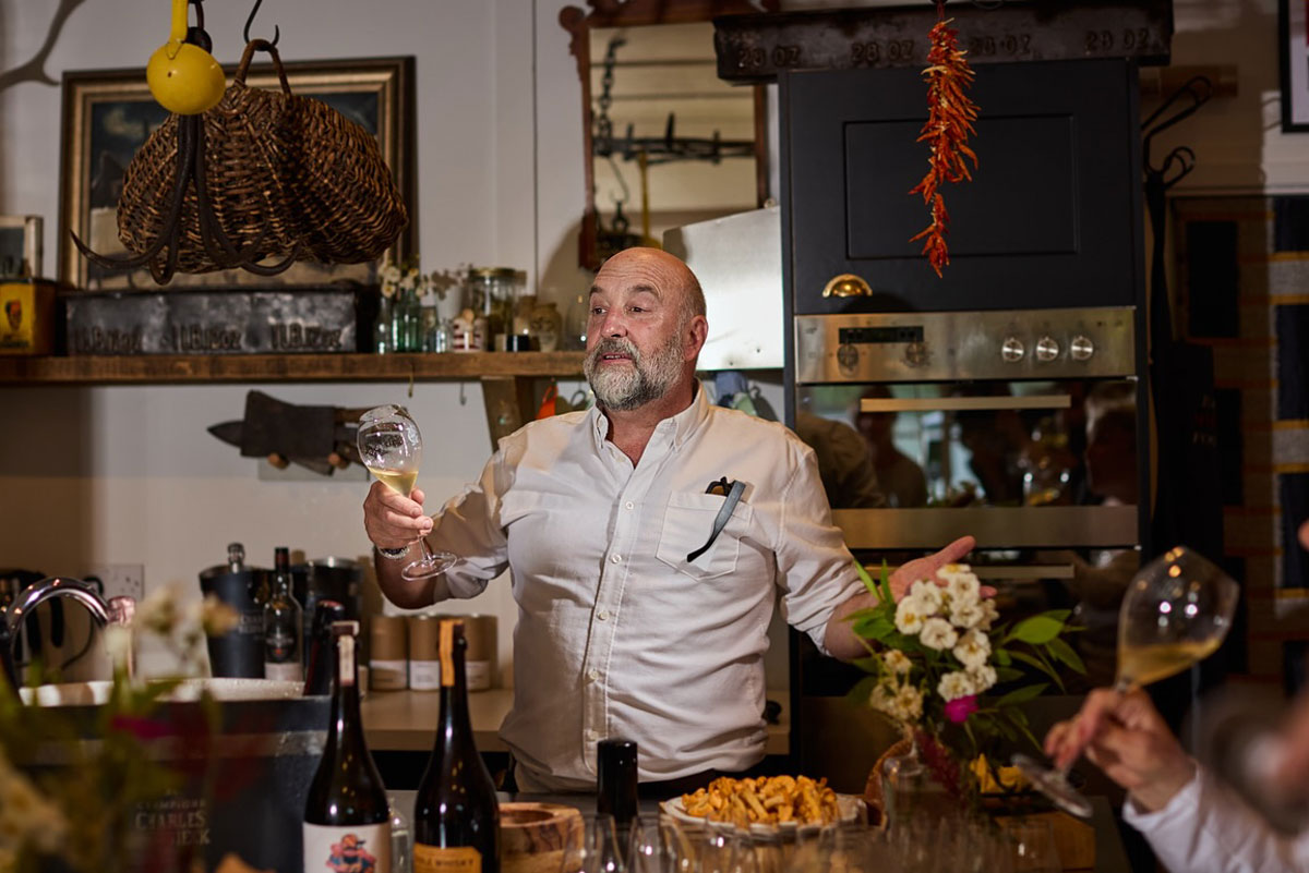 a jolly-looking bearded man holds forth over a table of food and wine