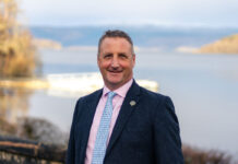 a well dressed man stands outdoors, with a Scottish loch behind him