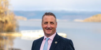 a well dressed man stands outdoors, with a Scottish loch behind him