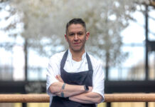 A man stands in chef's whites in front of a large window out to trees