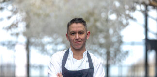 A man stands in chef's whites in front of a large window out to trees