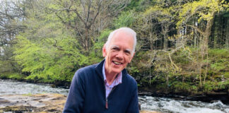 A distinguished gentleman sits smiling by a Scottish river
