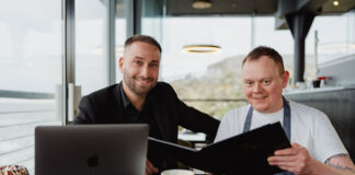 A well dressed man and a chef sit in a seafront restaurant