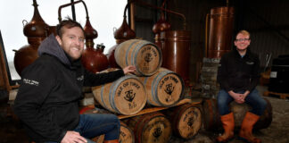 Two young men sit smiling beside a pile of whisky barrels