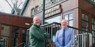 Two smiling men stand outside a Highlands hotel on a sunny day