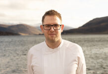 A young man stands in chef's whites outdoors on the shore of a Scottish sea loch