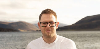 A young man stands in chef's whites outdoors on the shore of a Scottish sea loch