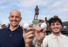 Two men stand holding golf balls, in front of a statue of Rabbie Burns