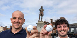Two men stand holding golf balls, in front of a statue of Rabbie Burns